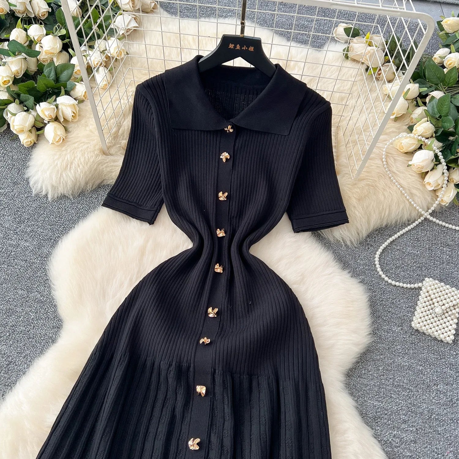 Black dress with gold buttons on a hanger against a decorative background with flowers and a rug.