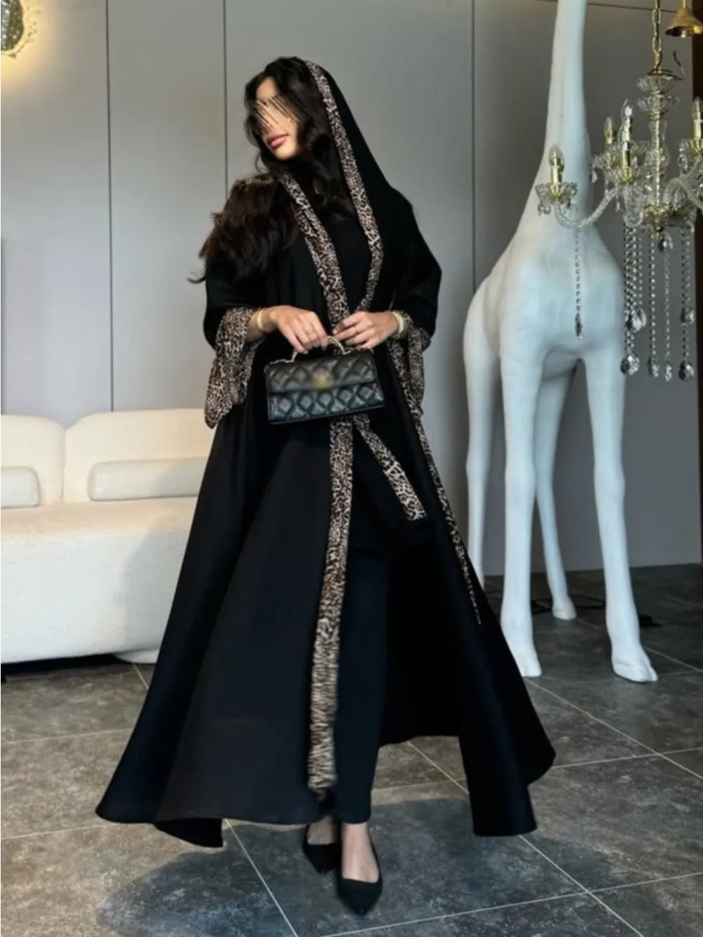 Person wearing a long black coat with decorative trim in a room with a white deer statue and chandelier.